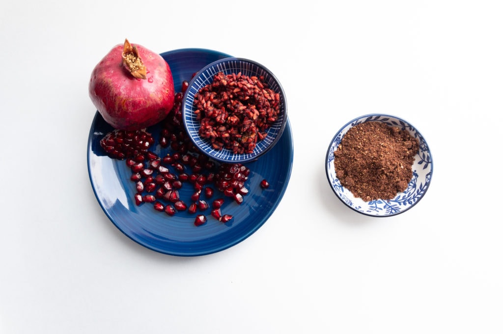 Flat lay of ground pomegranate seeds spice in bowl on white background with garnet fruit and berries in blue plate